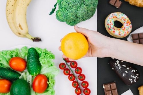 Photographie d'une personne tenant une orange, avec en fond deux côtés, blanc pour la nourriture saine avec avocat brocoli et tomates, et noir pour la nourriture transformée comme un donut, une barre de chocolat, un croissant...