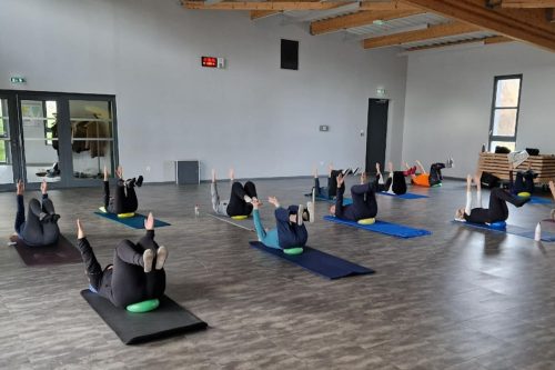 Photographie d'un groupe de l'activité Pilates en intérieur sur des tapis de yoga, sur le dos, bras et jambes en l'air