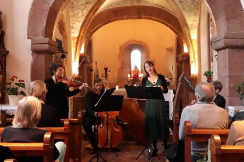 Photographie d'un concert se tenant dans une église, le groupe joue du violon, du violoncelle et de la basse, et une chanteuse se tient devant un micro