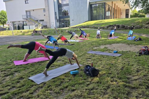 Photographie de l'activité yoga en extérieur, plusieurs participants sur des tapis en train de faire une variante du Dandayamana Bibhaktapada Janushirasana.